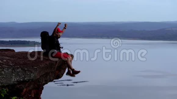 一个快乐的徒步旅行者坐在悬崖边上在一座岩山上举起双臂视频的预览图