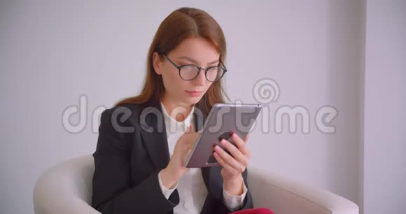 一位戴眼镜的年轻白种人女商人的特写肖像他在平板电脑上发短信坐在白色的扶手椅上视频的预览图