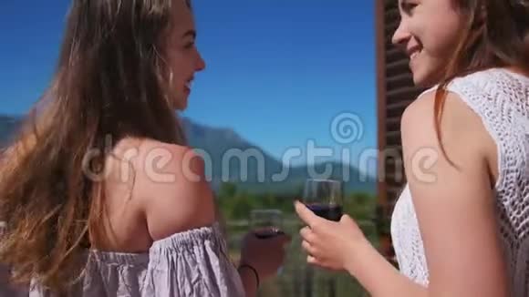 两位年轻女子站在阳台上喝着红酒欣赏着山和森林的景色视频的预览图
