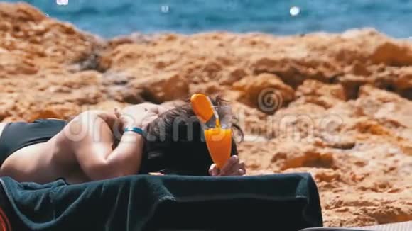 美丽的女孩躺在海滩上的休息室里手里拿着一个鸡尾酒视频的预览图