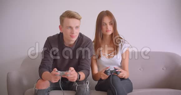 一对年轻可爱的白种人游戏玩家夫妇坐在室内的沙发上玩电子游戏的特写镜头视频的预览图