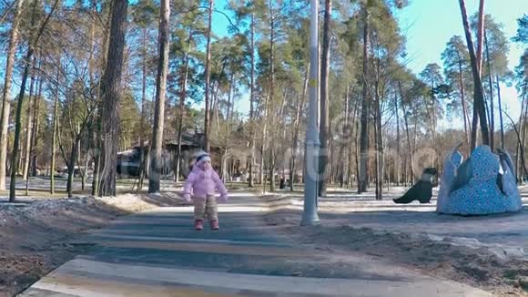 一个穿着粉红色夹克和条纹帽子的小女孩在妈妈的怀抱和微笑中沿着春天公园小路跑视频的预览图