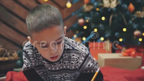 可爱的男孩穿着节日礼服躺在地板上写信给圣诞老人圣诞树上有灯和礼物视频的预览图