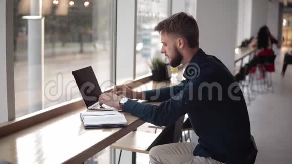 年轻英俊的商人坐在coferoom的笔记本前开始使用智能手机与他交谈视频的预览图