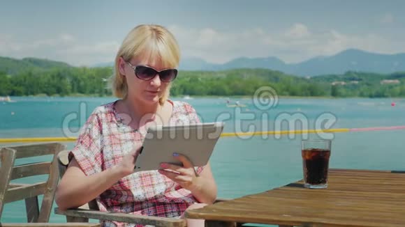 假期的技术一位拿着平板电脑的女士坐在湖和山背景的咖啡馆里的桌子旁视频的预览图