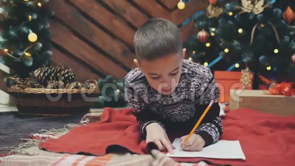 可爱的男孩穿着节日礼服躺在地板上写信给圣诞老人圣诞树上有灯和礼物视频的预览图