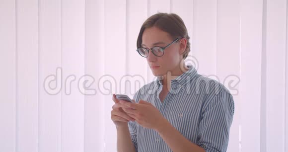 戴着眼镜戴着手机站在办公室室内的年轻白种人商人的特写照片视频的预览图