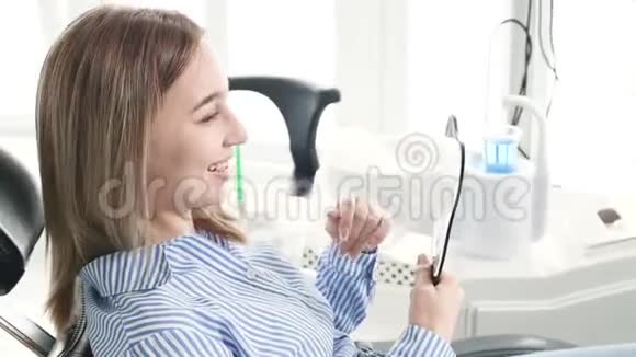 一个微笑的金发女孩在牙科椅上的肖像她的手里拿着一面牙齿形状的镜子快乐顾客牙科视频的预览图