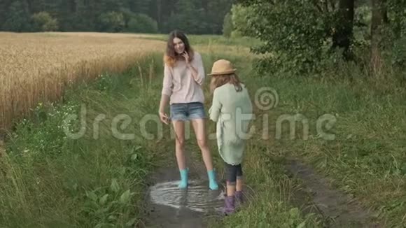 两个穿着靴子的快乐女孩姐妹在乡间小路上的雨水中嬉戏视频的预览图