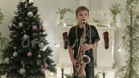 一个英俊的男孩在圣诞树的背景下演奏萨克斯风假日家庭音乐会视频的预览图