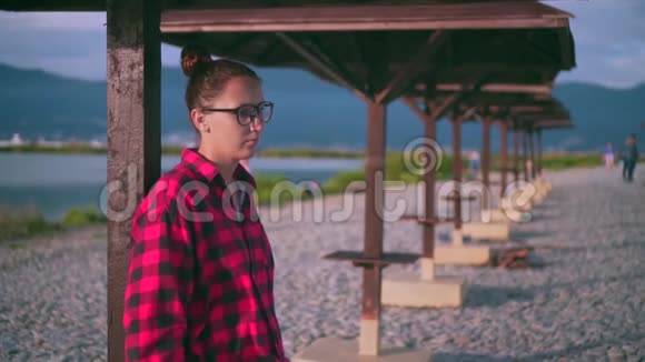 漂亮的年轻女孩带着眼镜和红色格子衬衫放松背景海滩上有许多木制阳伞视频的预览图