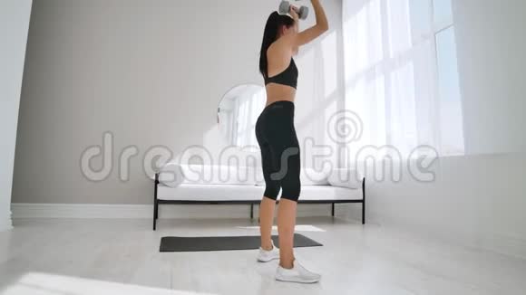 阳光明媚的一天一个穿着黑色运动服的年轻黑发女孩在一间白色公寓里用哑铃做蹲慢慢视频的预览图