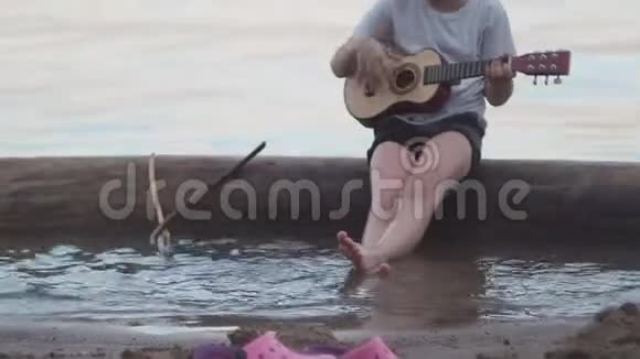 坐在海里的木头上弹吉他的小男孩他微笑着唱着快乐的歌视频的预览图