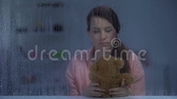 无助的女人受伤的脸在雨窗后拥抱玩具熊视频的预览图