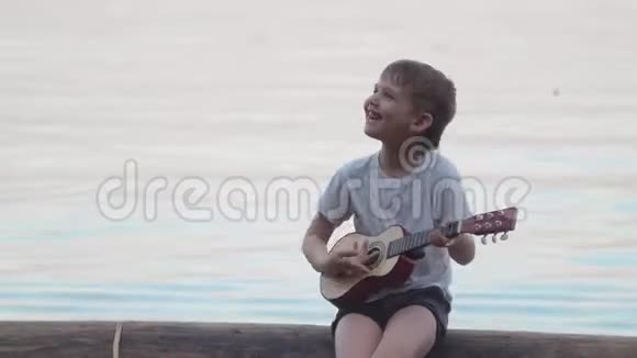坐在海里的木头上弹吉他的小男孩他微笑着唱着快乐的歌视频的预览图
