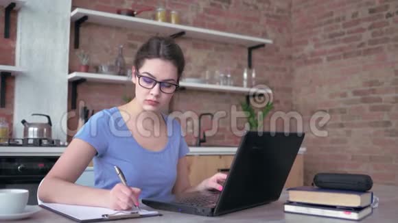 戴眼镜的年轻女子在家里的笔记本电脑上工作或学习时在剪贴板上写笔记视频的预览图