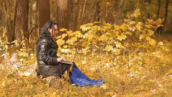 一个穿着蓝色靴子的黑发女人坐在秋天的公园里用笔记本电脑工作视频的预览图