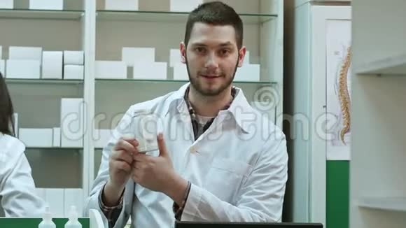 年轻的malepharmacist手里拿着一瓶白色的空白药丸在他的同事工作的时候推销药物视频的预览图