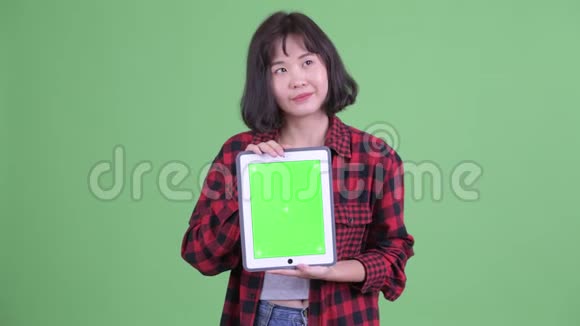 快乐的亚洲时髦女人一边思考一边展示数码平板电脑视频的预览图