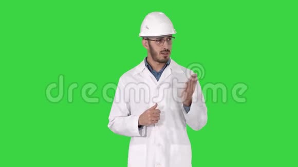 身着白色长袍和头盔的科学家用手指着侧面向绿色屏幕上的摄像机解释视频的预览图