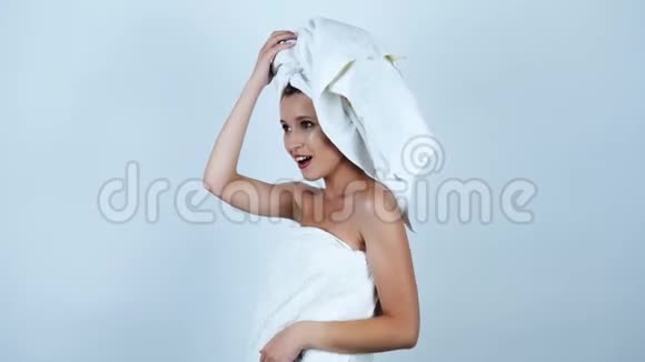 年轻漂亮的女人头上戴着白毛巾身体上裹着白毛巾双臂向上跳舞在一个孤立的白色酒吧里感到快乐视频的预览图