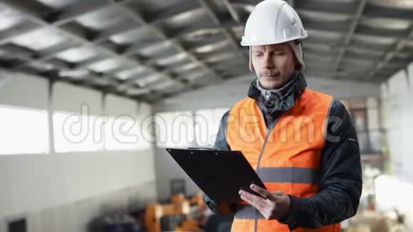 戴着安全帽穿着工作服的男人胡须和胡子站在机库的中央注意到有平板电脑的工具视频的预览图