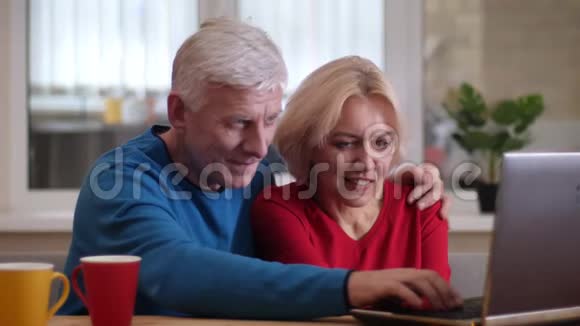 一对可爱的老夫妇在办公桌上的笔记本电脑上在一个舒适的房间里用杯子和茶来浏览特写镜头视频的预览图