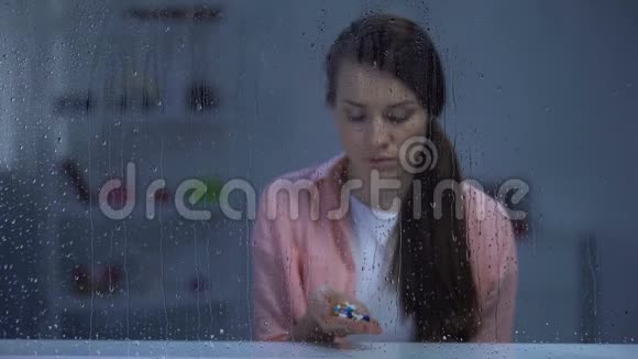 沮丧的女人在雨窗后的手掌上拿着药丸昂贵的药视频的预览图