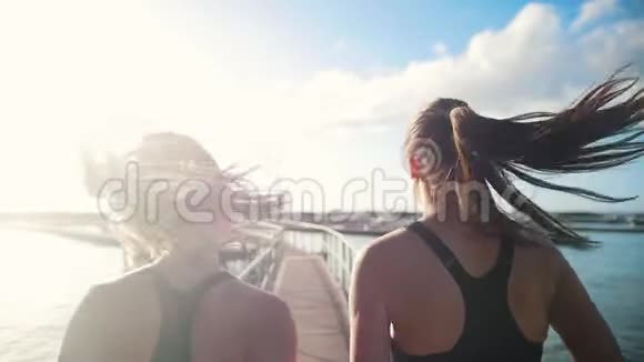 两个年轻女子在阳光明媚的日子里在桥上向海港慢跑视频的预览图