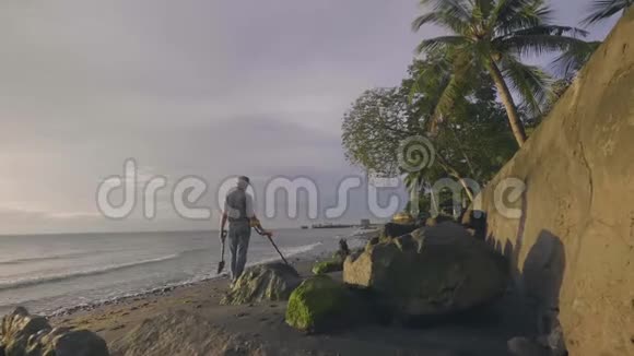 人类正在用金属探测器扫描海滩并在日落时与他的狗在海洋附近散步视频的预览图