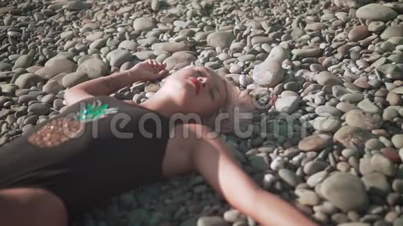穿着泳衣躺在岩石海岸上的年轻性感女孩金发碧眼的一件泳衣躺在海滩的石头上视频的预览图