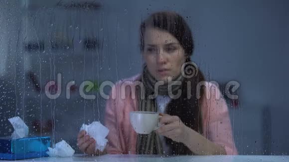 戴着围巾的患病女士在雨天在家打喷嚏和喝热茶视频的预览图