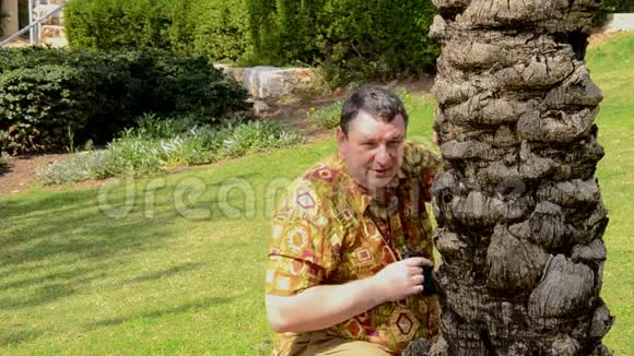 穿着阿洛哈衫的成熟男人透过望远镜看公园里的棕榈树干视频的预览图