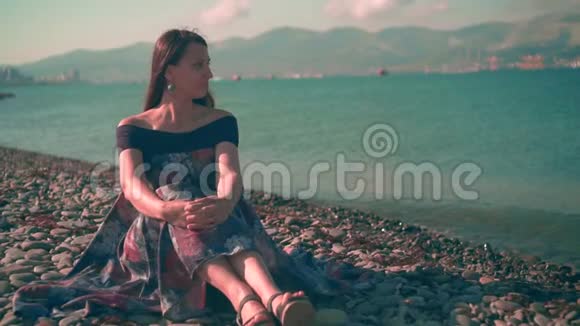 美丽的女孩穿着五颜六色的裙子坐在海滩上女孩的身后是大海和山脉视频的预览图