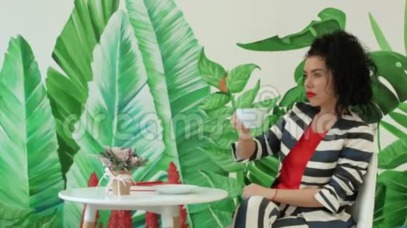 一个穿着条纹西装的年轻漂亮女人正在绿叶背景下喝咖啡视频的预览图