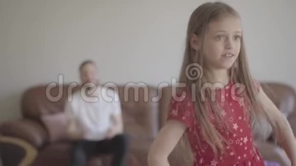 可爱的小滑稽女孩穿着红色连衣裙在蓬松的地毯上的前景中跳舞的肖像坐在那儿的父亲视频的预览图