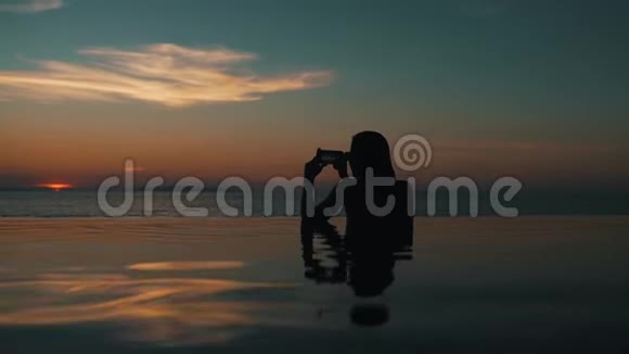 坐在游泳池里的女孩在海洋上拍摄日落视频的预览图