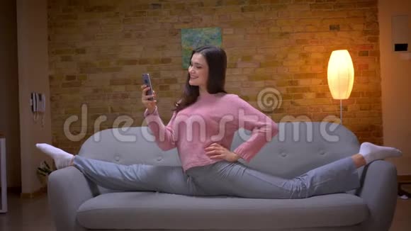 一位年轻漂亮的褐发白种人女性在舒适的沙发上用电话和麻绳坐着的特写镜头视频的预览图