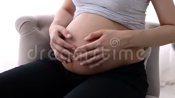 年轻的欧洲孕妇坐在白色背景的扶手椅上轻轻抚摸着她赤裸的大肚子视频的预览图