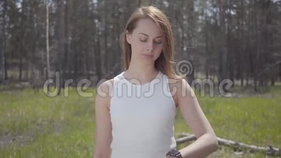 一位美丽的年轻女子棕色长发看着手表在绿色的空地上等着某人视频的预览图