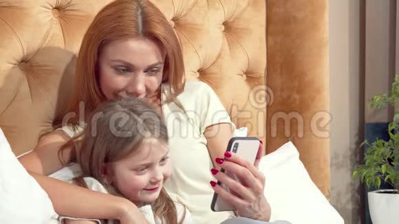 美丽的女人和她可爱的小女儿一起在家里用智能手机视频的预览图