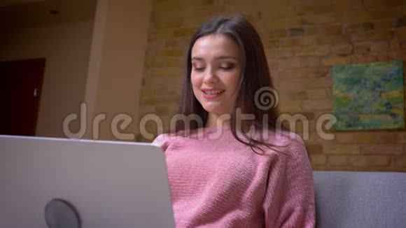 年轻漂亮的棕色白种人女学生用笔记本电脑坐在舒适的沙发上的特写镜头视频的预览图