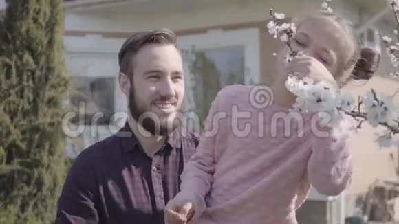 肖像年轻的大胡子父亲举起他的小微笑的女儿这样她就能闻到树上的花朵家庭视频的预览图