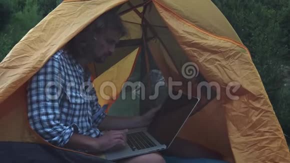自由职业者使用笔记本电脑坐在森林的露营帐篷从事新启动项目的自由职业者视频的预览图