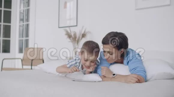 在白色的卧室里妈妈和儿子看着平板电脑笑了起来快乐的一家人在床上晨读一本书视频的预览图