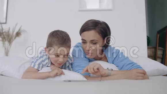 快乐的年轻母亲在她蹒跚学步的儿子睡觉前在数码平板上展示卡通快乐的妈妈视频的预览图