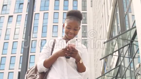 年轻快乐的非洲女学生穿着白色基本衬衫背着皮背包看着她的智能手机视频的预览图