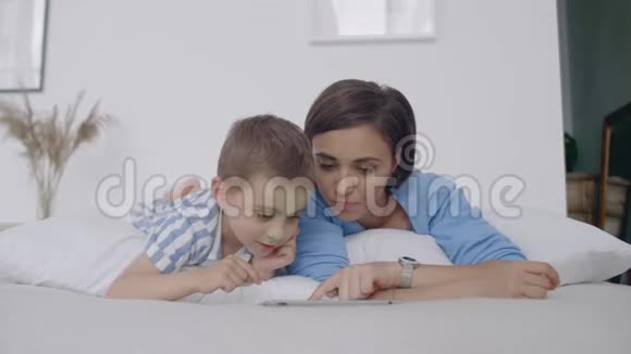 在白色的卧室里妈妈和儿子看着平板电脑笑了起来快乐的一家人在床上晨读一本书视频的预览图