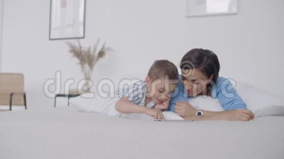 母子俩看着躺在白色床上的平板屏幕在平板电脑上和你儿子玩游戏视频的预览图