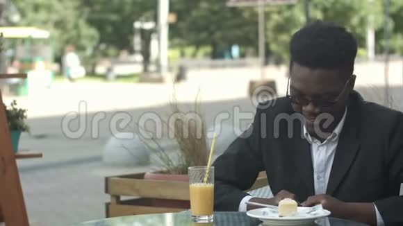 放松无忧无虑的年轻美国黑人男子戴着时尚眼镜独自坐在咖啡桌上使用智能手机阅读文字视频的预览图